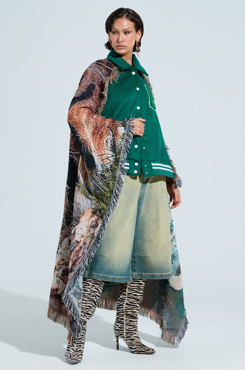 A person stands confidently in an evergreen wool-blend jacket, knee-length denim skirt, and animal print boots, draped in the colorful FIRST ROUND DRAFT PICK TAPESTRY BOMBER adorned with a subtle tiger design against a plain background.