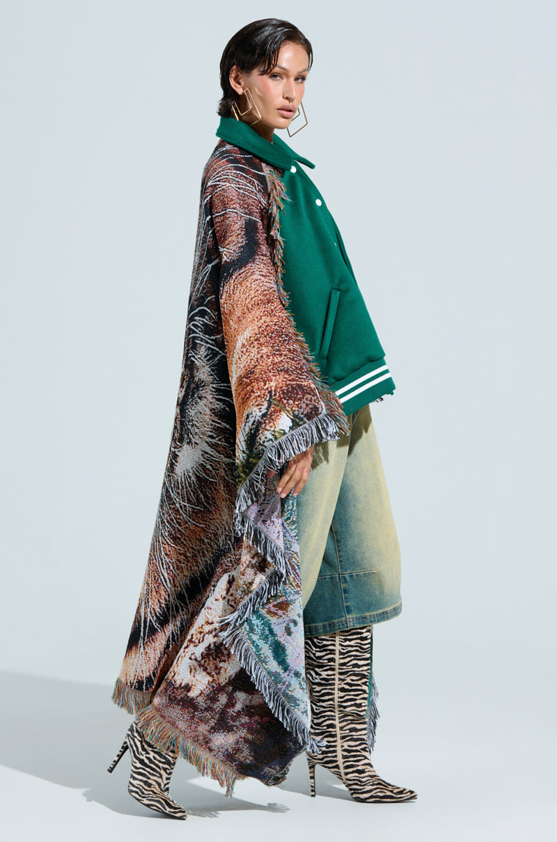 A model poses in a FIRST ROUND DRAFT PICK TAPESTRY BOMBER with a long, fringed cape featuring an abstract tiger design, paired with light-colored, wide-leg jeans and zebra-striped knee-high boots against a light gray background.
