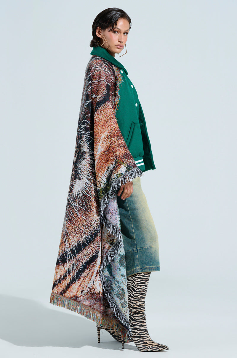 A person in a FIRST ROUND DRAFT PICK TAPESTRY BOMBER under an animal print cape stands in profile, wearing knee-length faded denim shorts and zebra-patterned knee-high boots. The plain background highlights the outfit's bold textures and patterns.