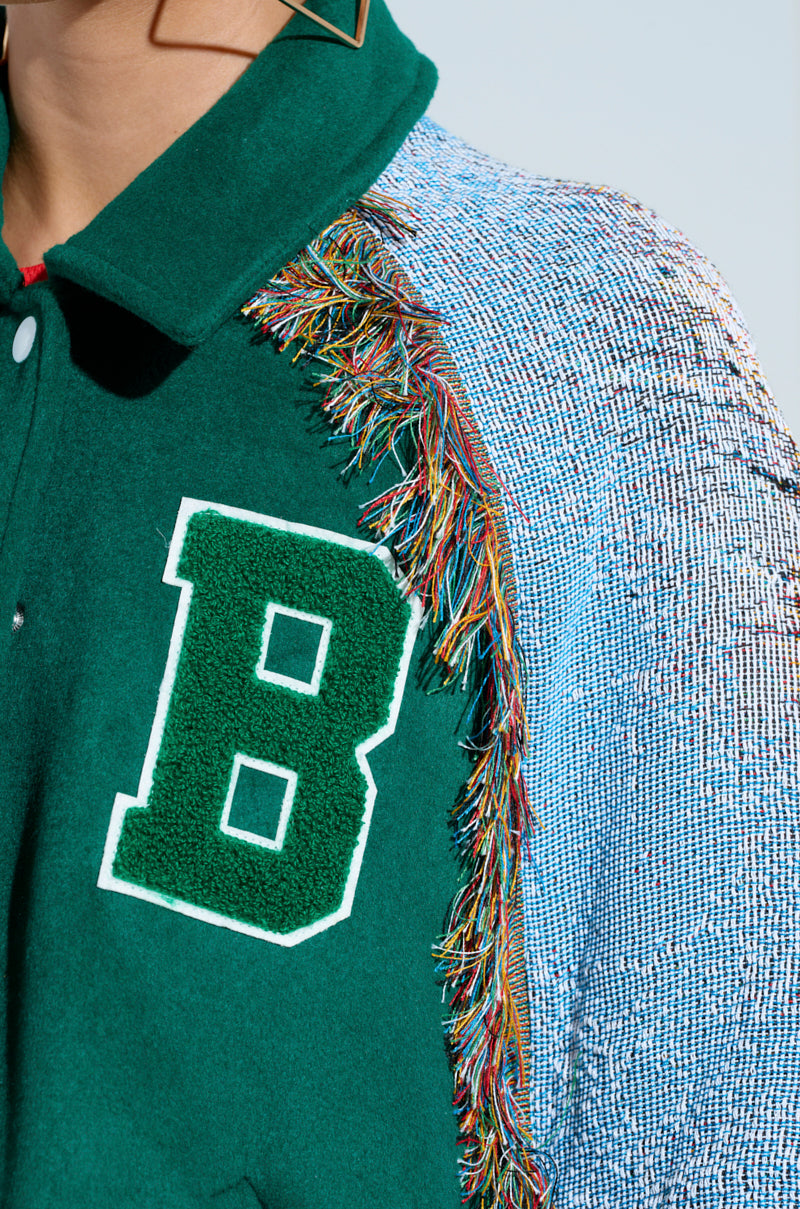 Close-up of the FIRST ROUND DRAFT PICK TAPESTRY BOMBER, a multicolored jacket with a large green "B" on the front. Made from an evergreen wool-blend, it features textured fabric and shoulder fringe, with blue, silver, and pink woven sleeve patterns.