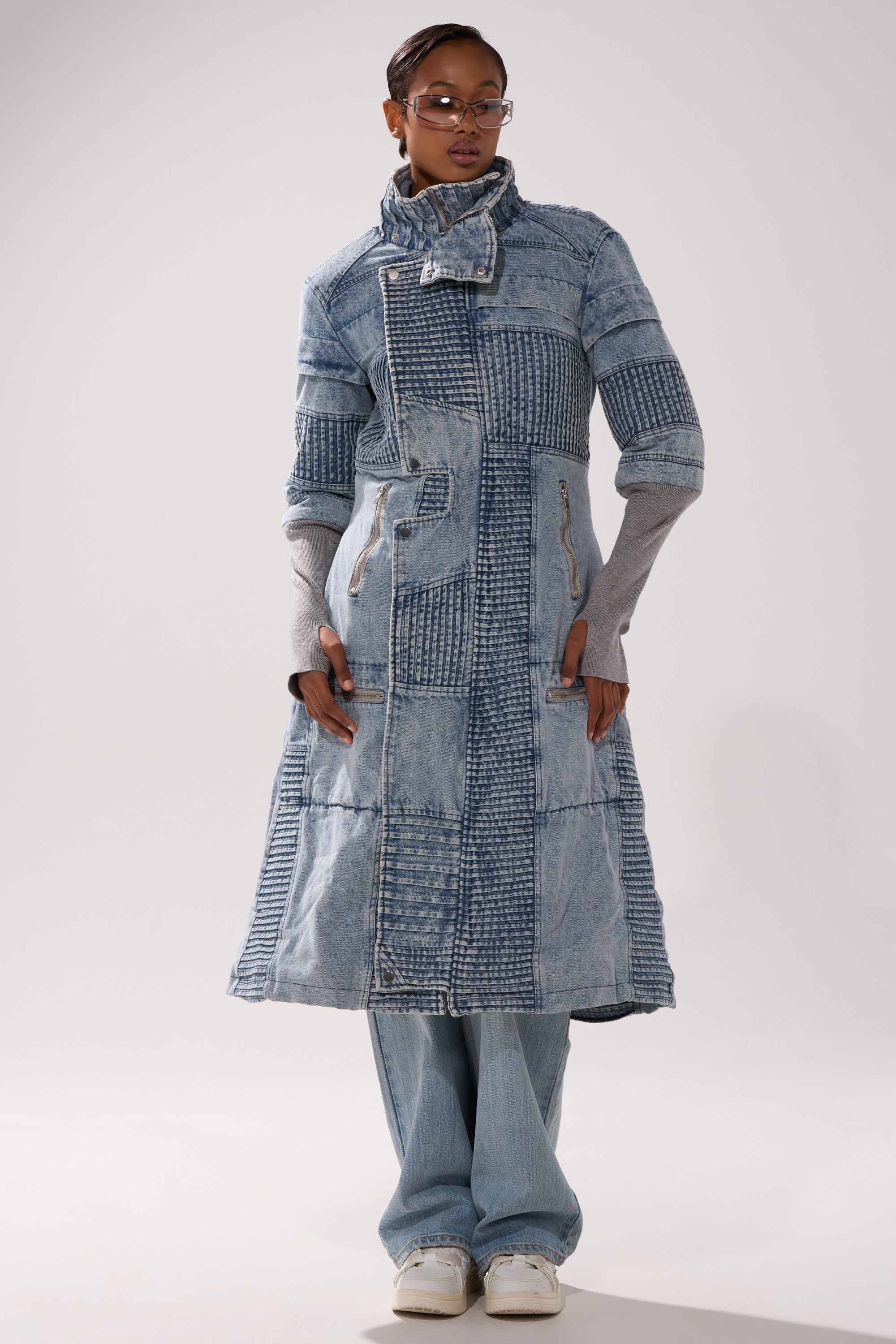 A person wearing glasses and the VORTEX DENIM TRENCH COAT IN BLUE with gray sleeves stands with hands in pockets, paired with light blue jeans and white sneakers against a plain background.