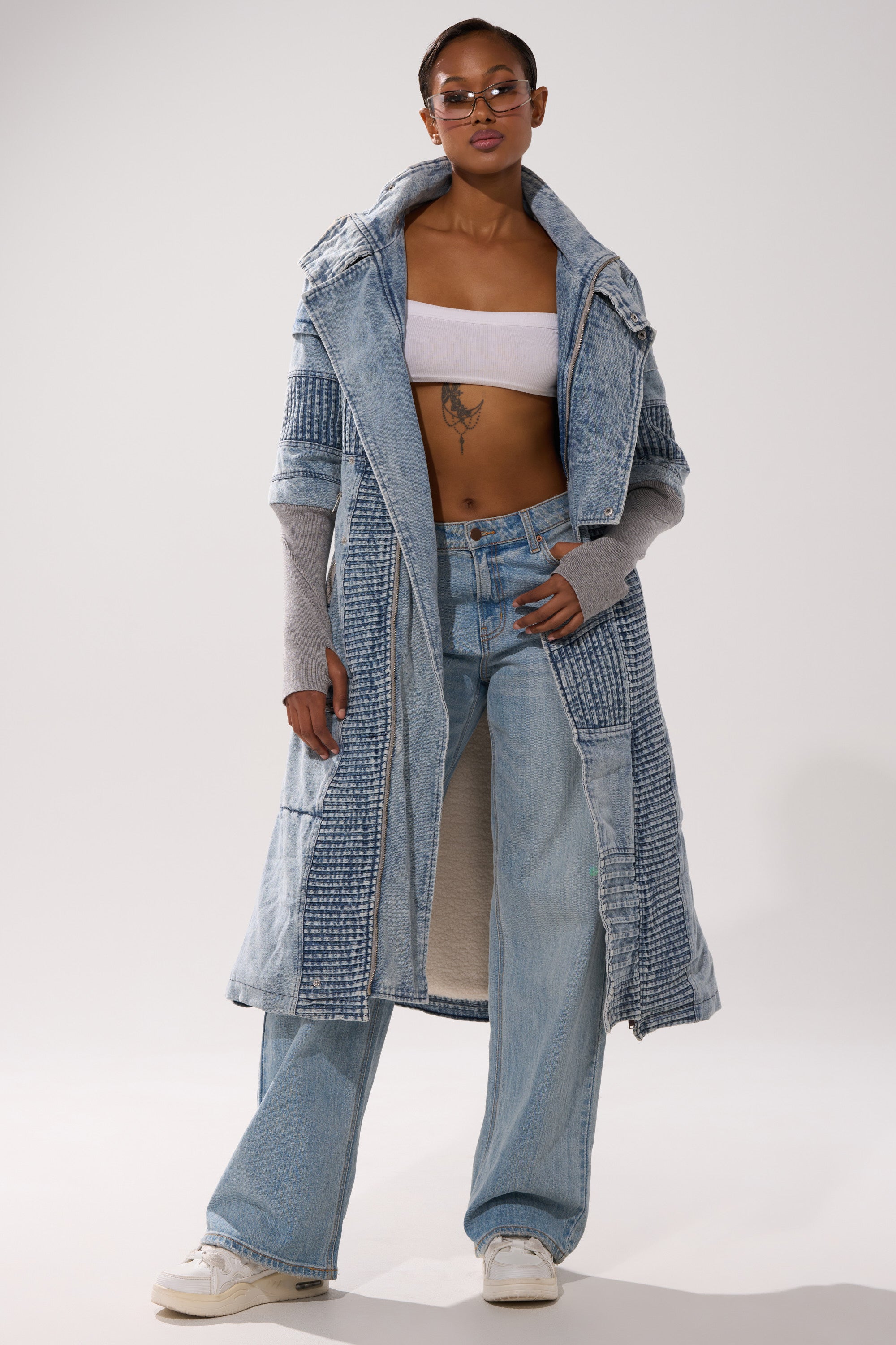 A woman in sunglasses, a white bandeau, loose jeans, white sneakers, and the VORTEX DENIM TRENCH COAT IN BLUE stands confidently against a plain background.