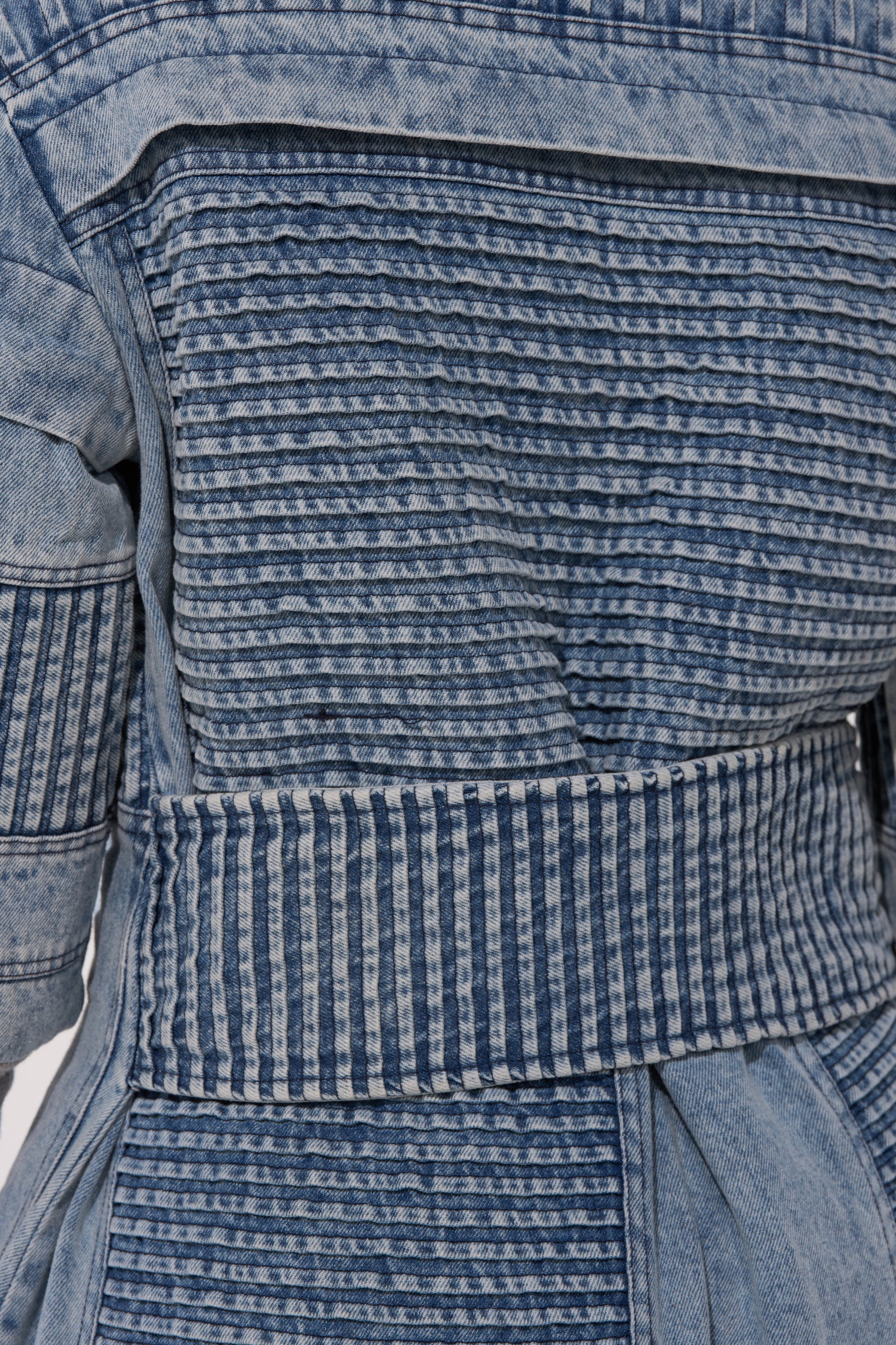 Close-up of the VORTEX DENIM TRENCH COAT IN BLUE, showcasing thick, structured denim with distinctive stitching, a belt-like section, and quilted textures created by horizontal and vertical lines.