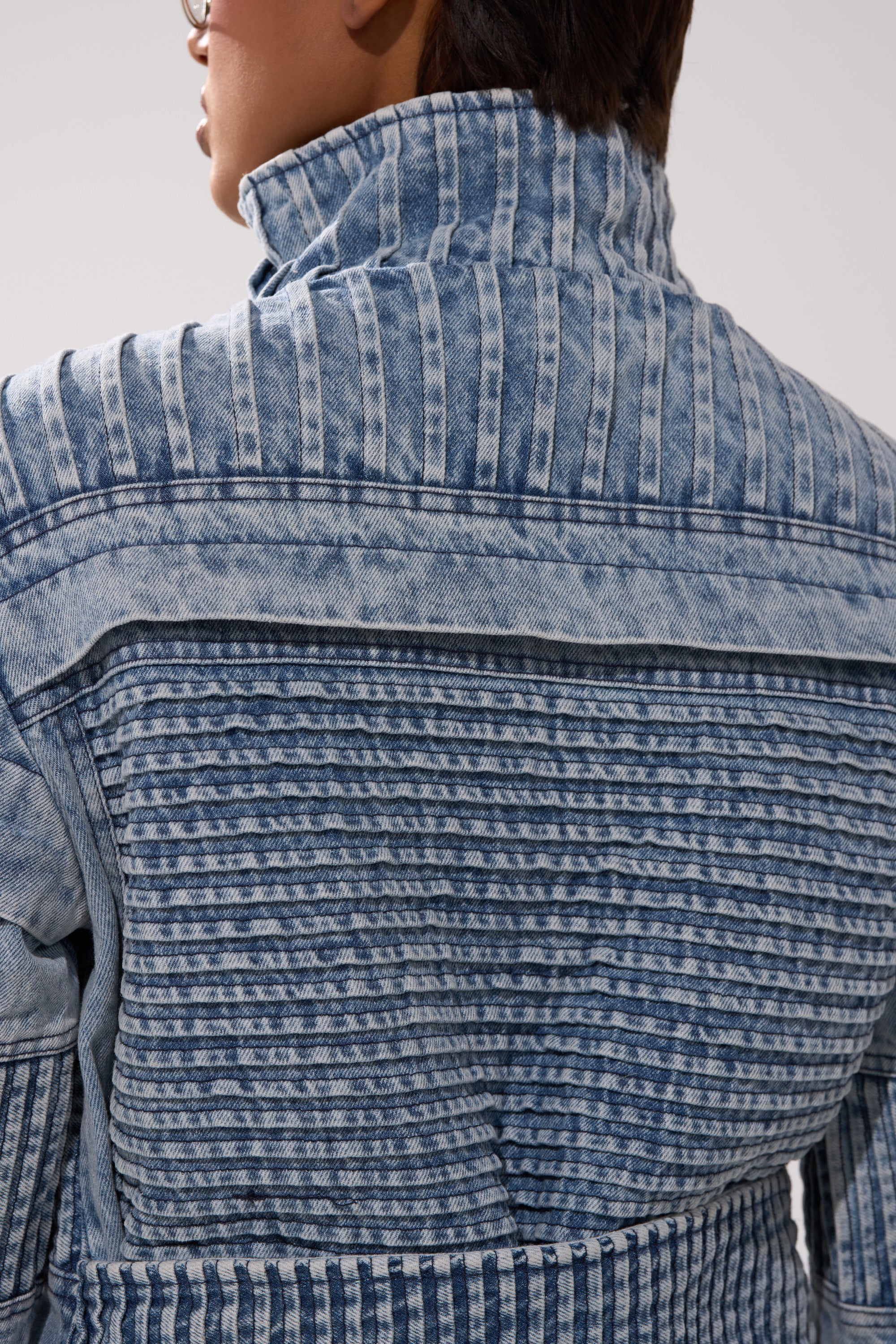 Close-up of a person from the back wearing the VORTEX DENIM TRENCH COAT IN BLUE, featuring vertical and horizontal striped textured patterns with the collar turned up.