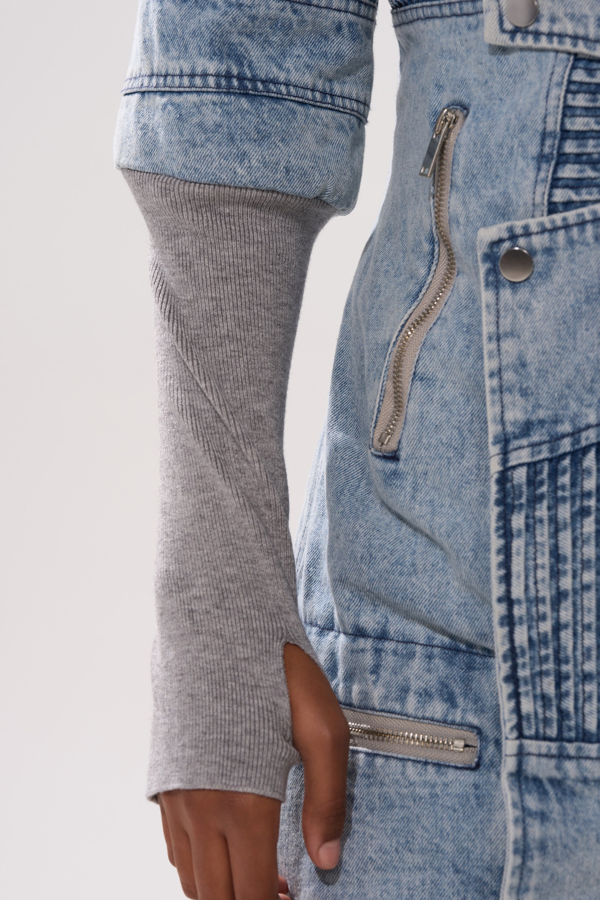A person wearing the VORTEX DENIM TRENCH COAT IN BLUE with zippers and a long gray knit sleeve featuring a thumb hole, revealing part of their hand.