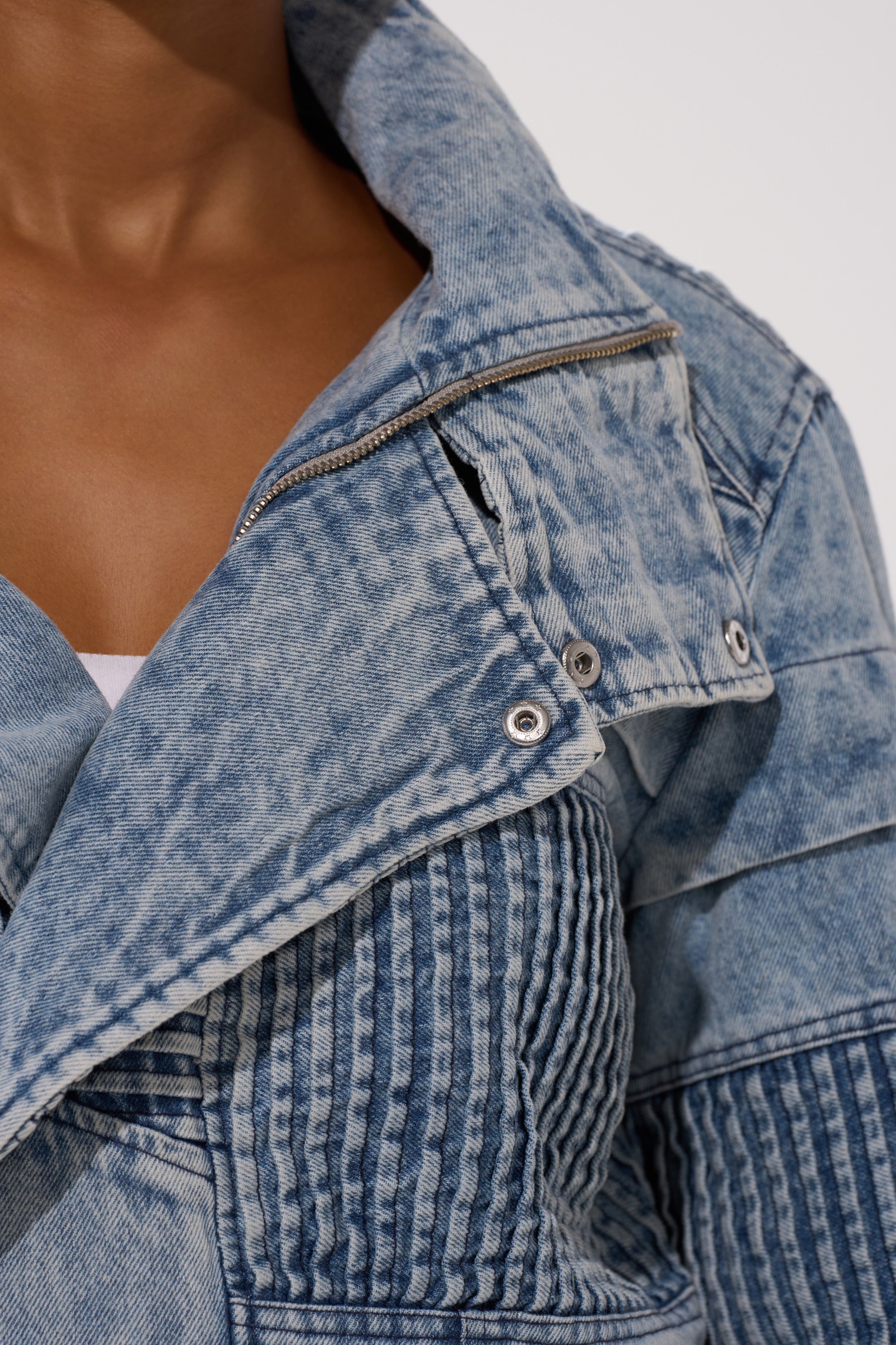 Close-up of a person wearing the VORTEX DENIM TRENCH COAT IN BLUE, featuring light blue acid-washed denim, metal buttons, and stitched details over a white top. The open collar reveals part of the neck and chest.