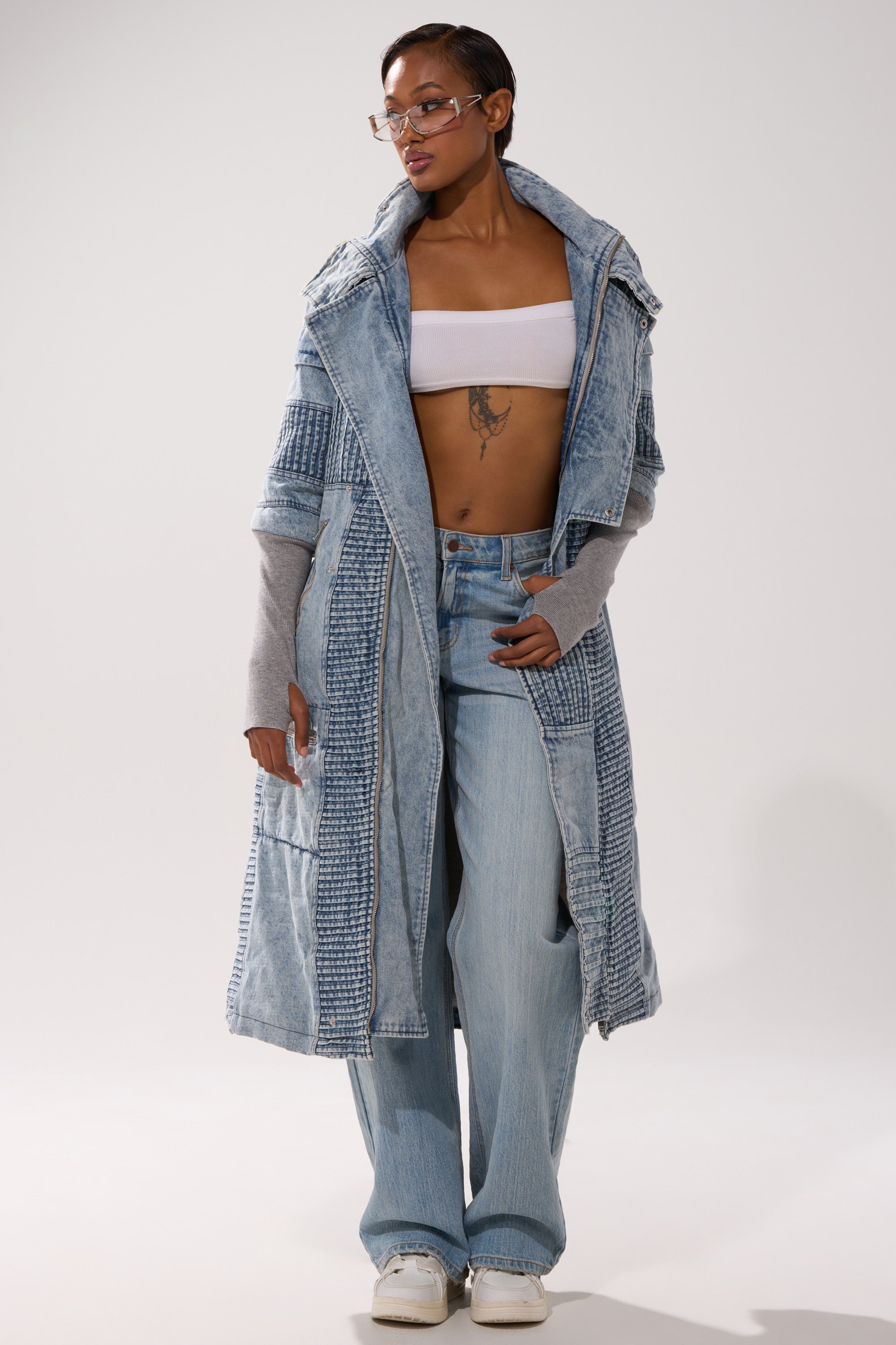 A person wearing the VORTEX DENIM TRENCH COAT IN BLUE over a white bandeau top, wide-leg light blue jeans, gray arm warmers, white sneakers, and clear glasses stands confidently against a plain background.