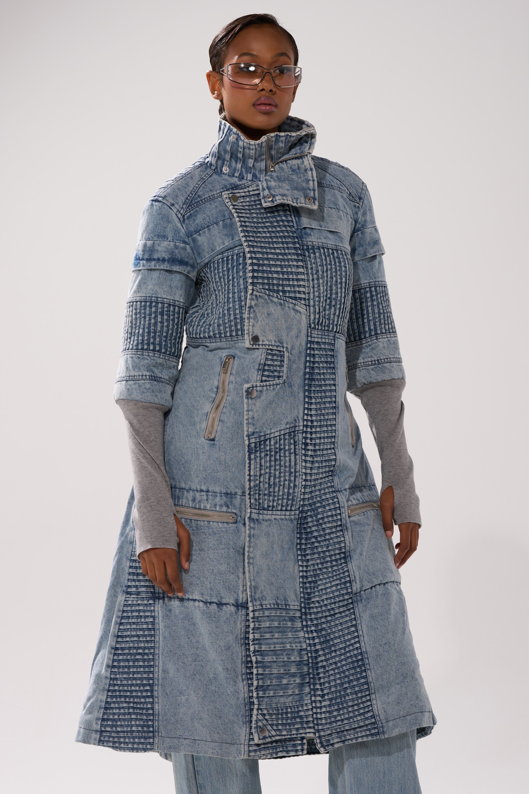 A person models the VORTEX DENIM TRENCH COAT IN BLUE, a long, high-collared patchwork denim coat with zipper and quilted details, layered over a gray long-sleeve top and blue jeans against a plain white background.