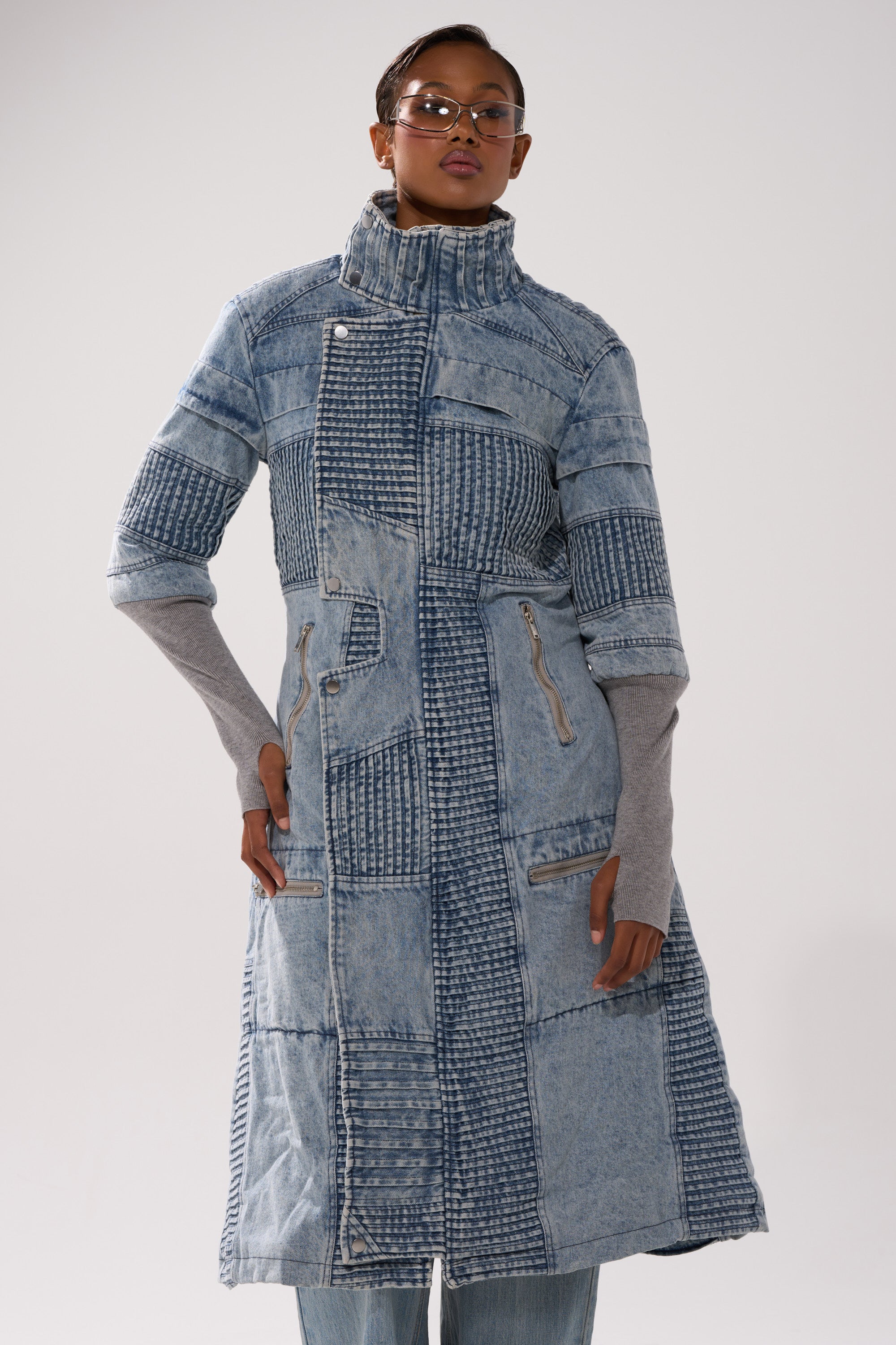 A woman wearing glasses and the VORTEX DENIM TRENCH COAT IN BLUE, layered over a gray long-sleeve top and blue jeans, poses against a plain light background.