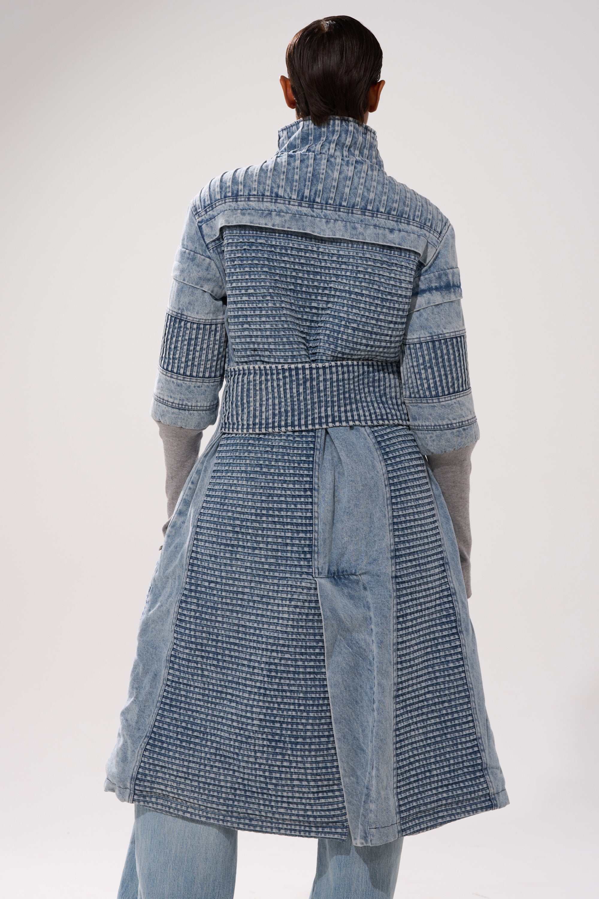 A person faces away from the camera, wearing the VORTEX DENIM TRENCH COAT IN BLUE— a long, textured denim coat with quilted and patterned panels— over light blue jeans, set against a plain light background.