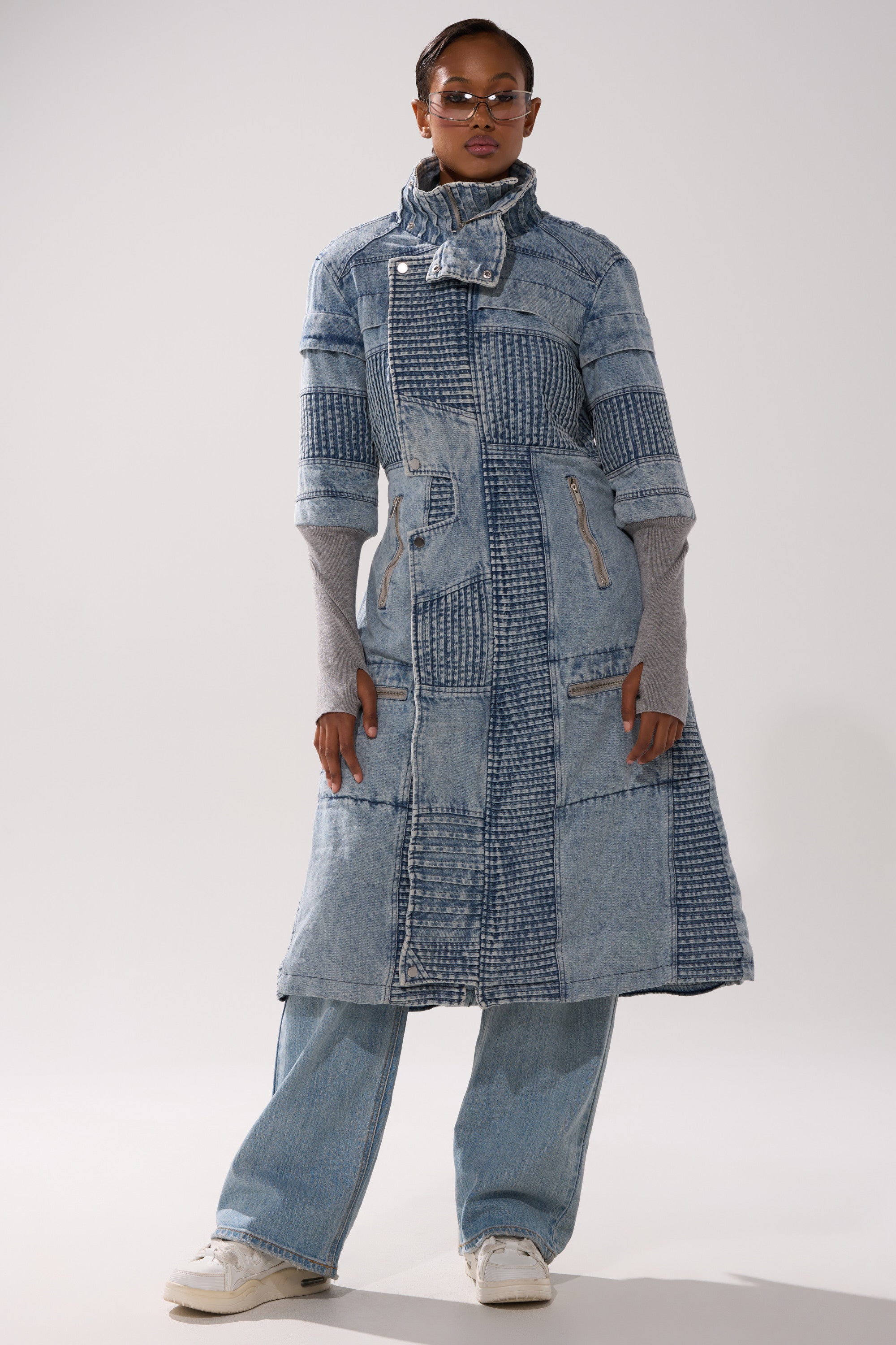 A person in the VORTEX DENIM TRENCH COAT IN BLUE, sunglasses, light blue jeans, and white sneakers stands against a plain light background.