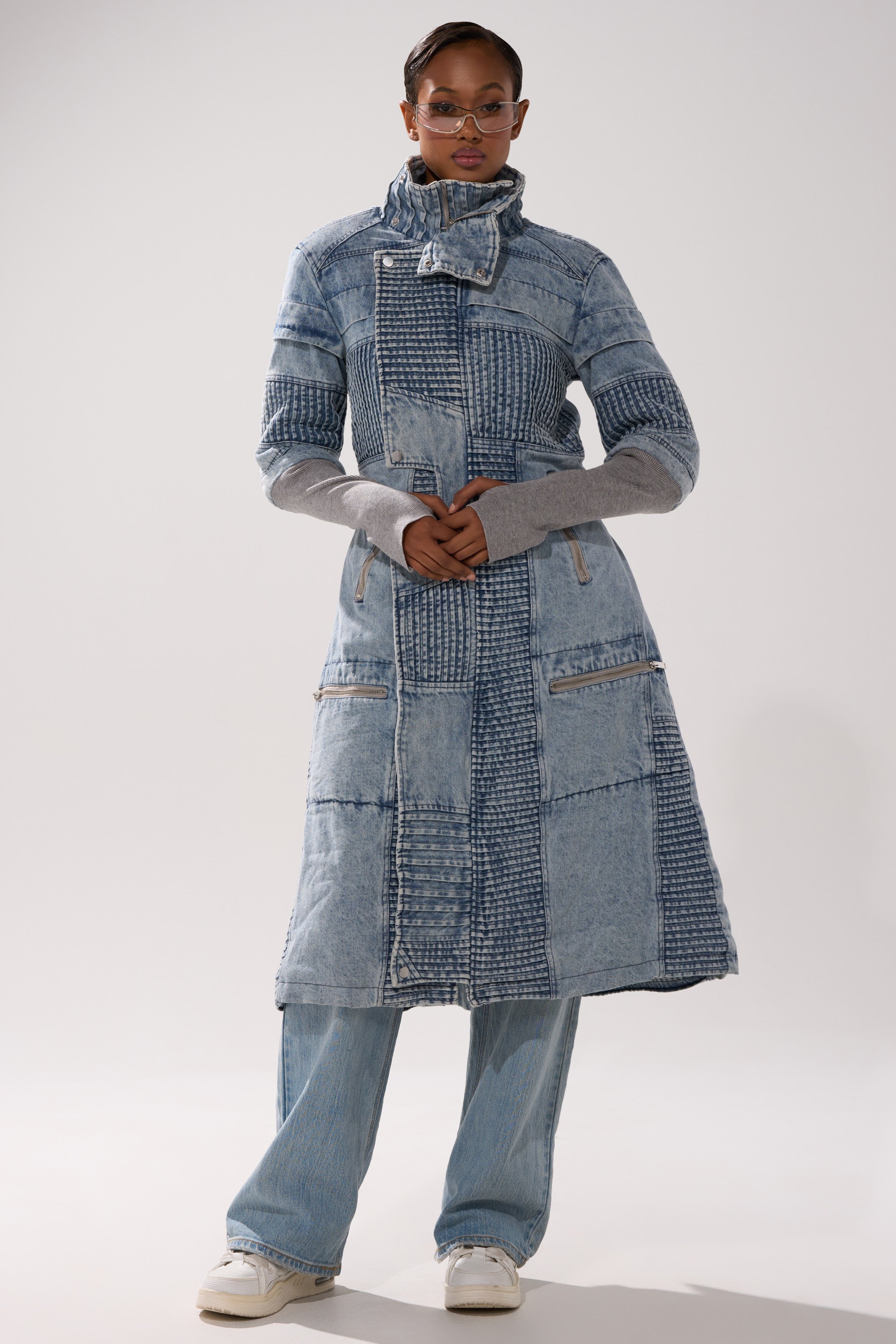 Wearing sunglasses and white shoes, a person stands against a plain light background in the VORTEX DENIM TRENCH COAT IN BLUE, layered over light blue jeans.