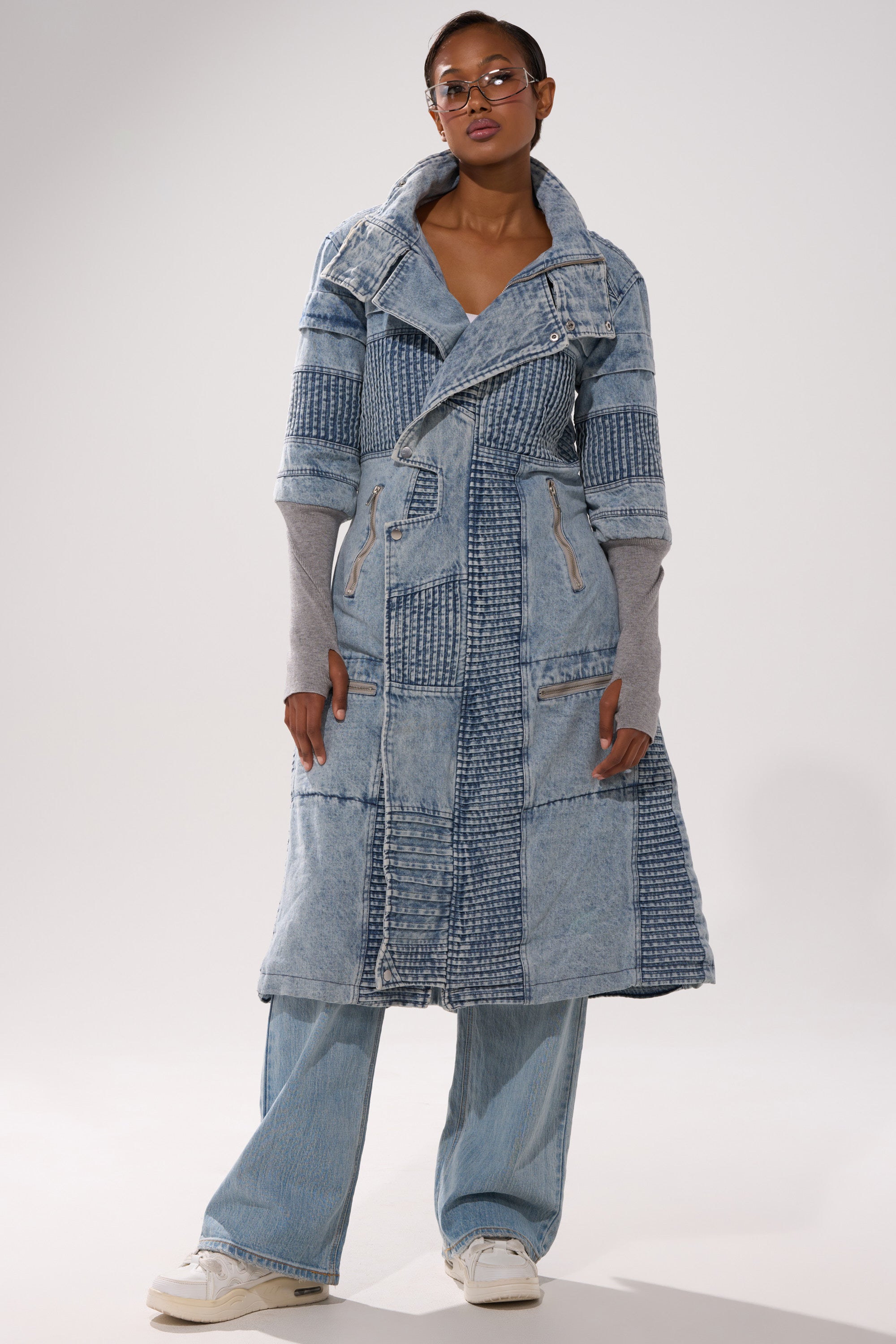 A person wears the VORTEX DENIM TRENCH COAT IN BLUE with light blue wide-leg jeans, a gray long-sleeve shirt, white sneakers, and glasses, standing against a plain light background.