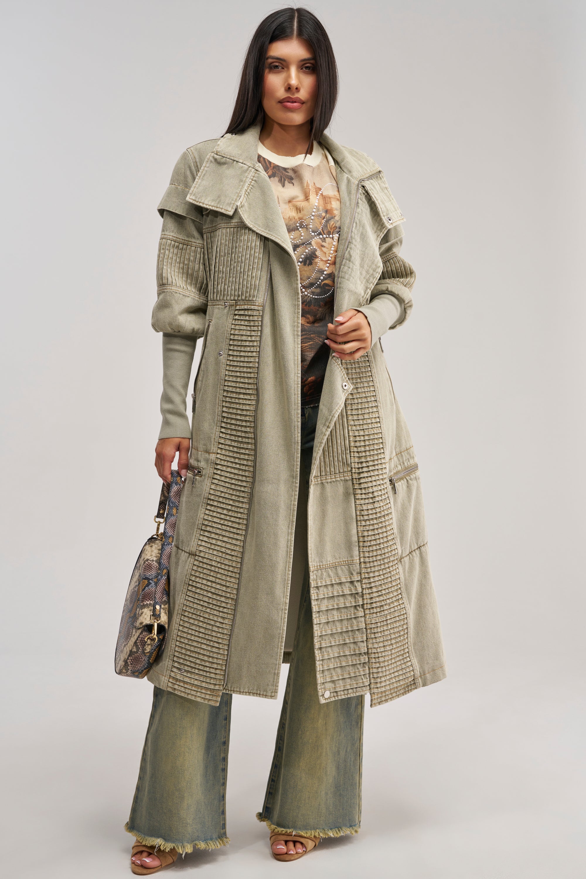 A woman with straight dark hair and a neutral expression faces forward wearing the VORTEX DENIM TRENCH COAT IN OLIVE, featuring a long, textured silhouette with faux sherpa lining. She styles it over a patterned top and wide-leg frayed jeans, holding a patterned handbag.