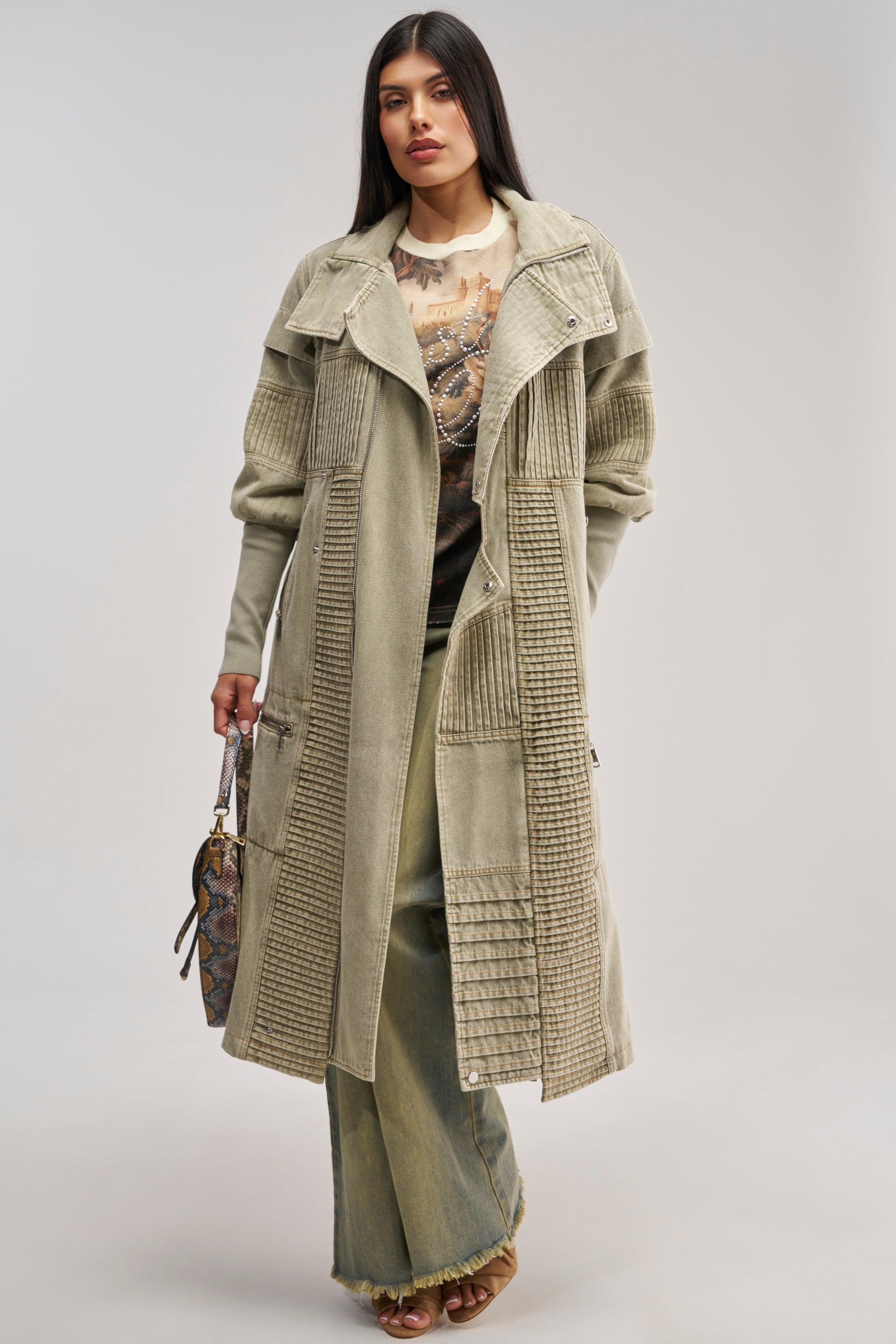A woman stands wearing the VORTEX DENIM TRENCH COAT IN OLIVE—a long, textured streetwear jacket with faux sherpa lining—layered over wide-leg pants and a printed top. She carries a patterned handbag, her straight dark hair framing her face as she looks confidently at the camera.