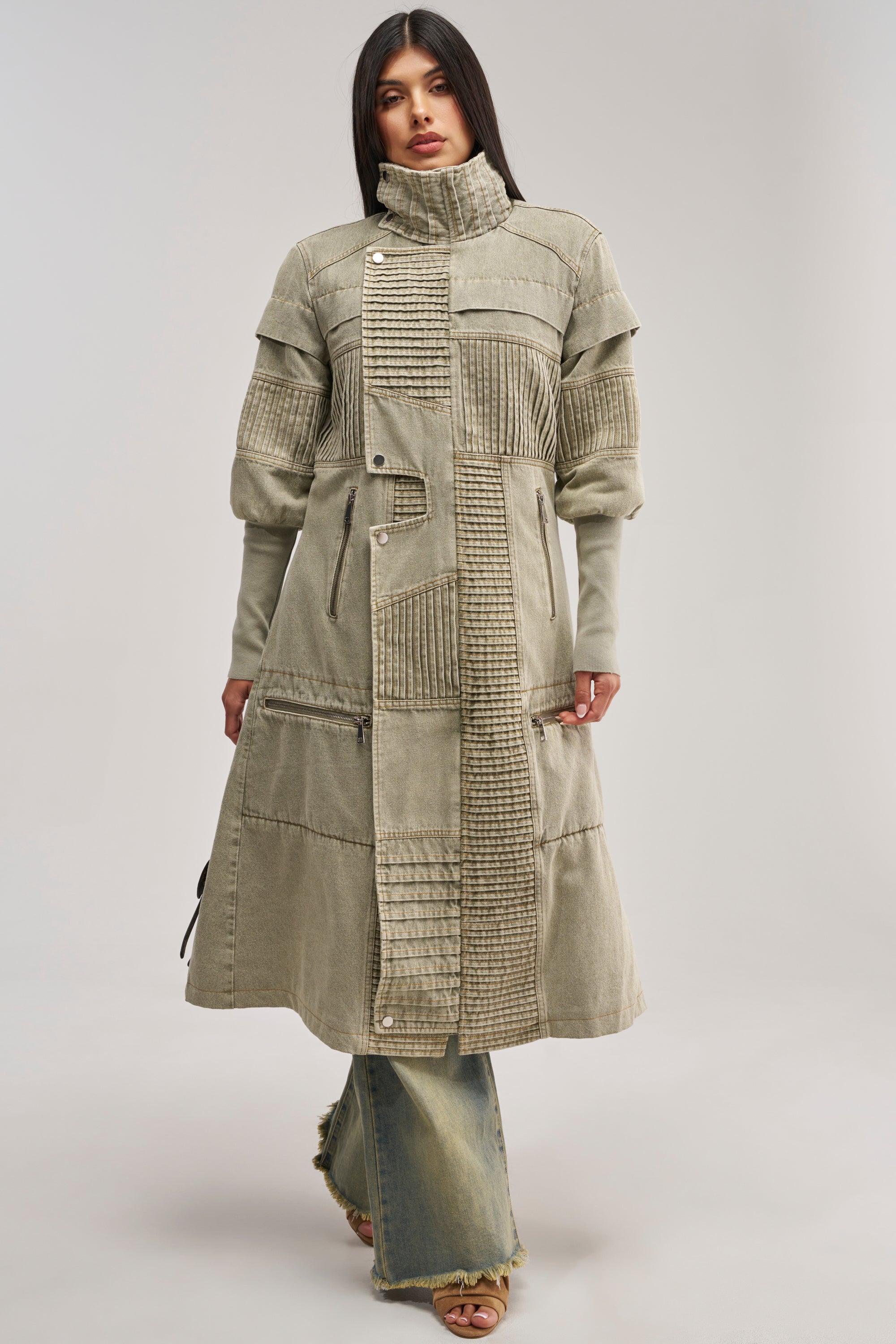 A woman stands against a plain background wearing the VORTEX DENIM TRENCH COAT IN OLIVE, a long, textured coat with patchwork details and faux sherpa lining, featuring a wide collar and asymmetrical buttons. She pairs it with frayed hem jeans and sandals.