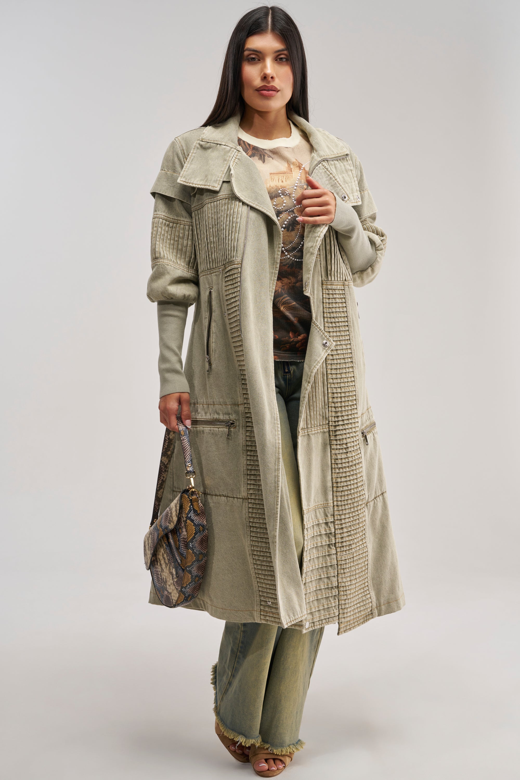 A woman wears the VORTEX DENIM TRENCH COAT IN OLIVE, a long, textured coat with faux sherpa lining, layered over a printed shirt and green jeans. She holds a snakeskin-patterned handbag and wears platform sandals, standing against a plain background.