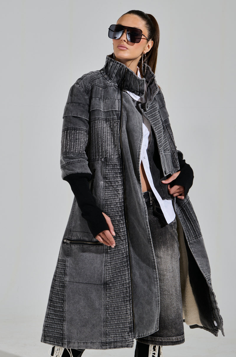 A person dons the VORTEX DENIM TRENCH COAT, a stylish gray knee-length coat featuring quilted detailing, ribbed patchwork, and black cuffs. They pair it with a white shirt and oversized sunglasses, their hair pulled back neatly.