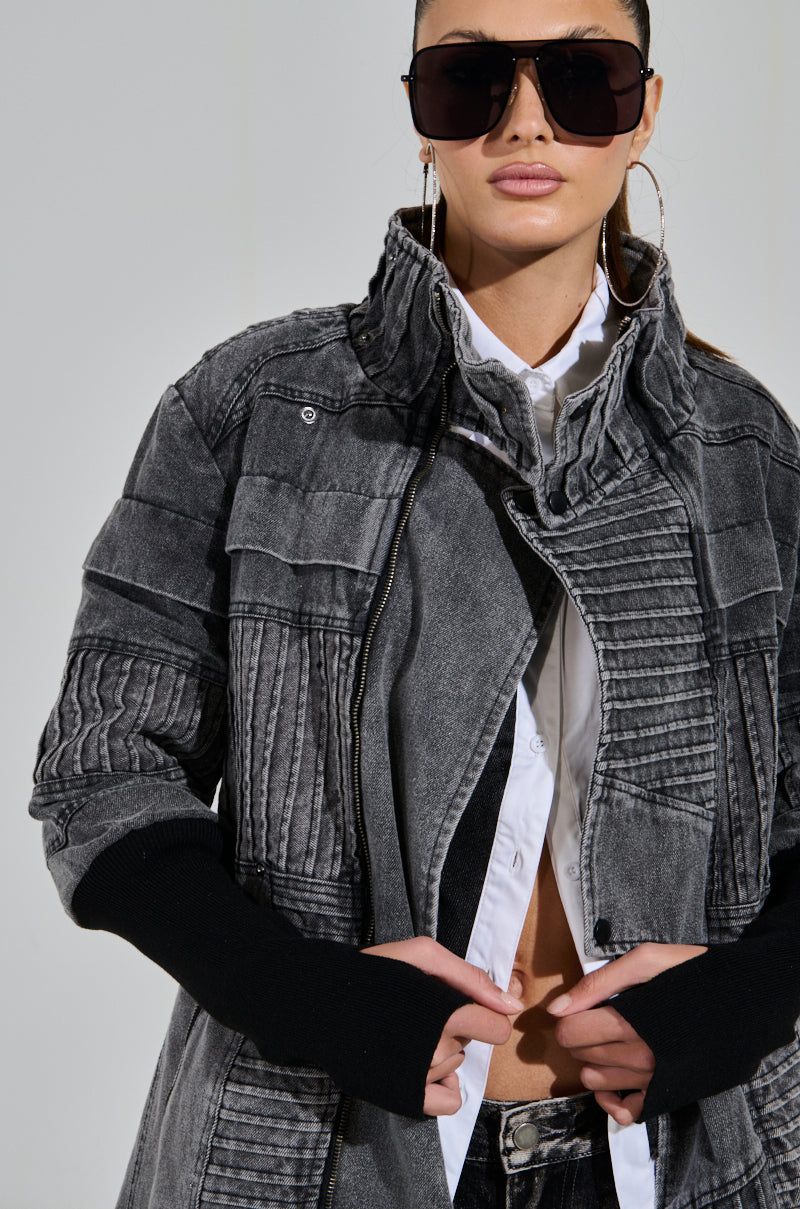 A person wearing oversized sunglasses and a textured grey VORTEX DENIM TRENCH COAT with ribbed patchwork detailing, along with a partially unbuttoned white shirt. They pose against a plain backdrop, hands adjusting the coat.