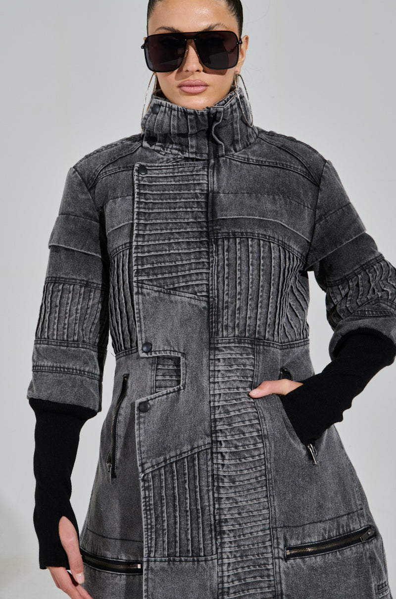 Someone confidently poses in a textured gray Vortex Denim Trench Coat with a high collar and ribbed patchwork detailing. They wear large sunglasses and have one hand in their pocket.