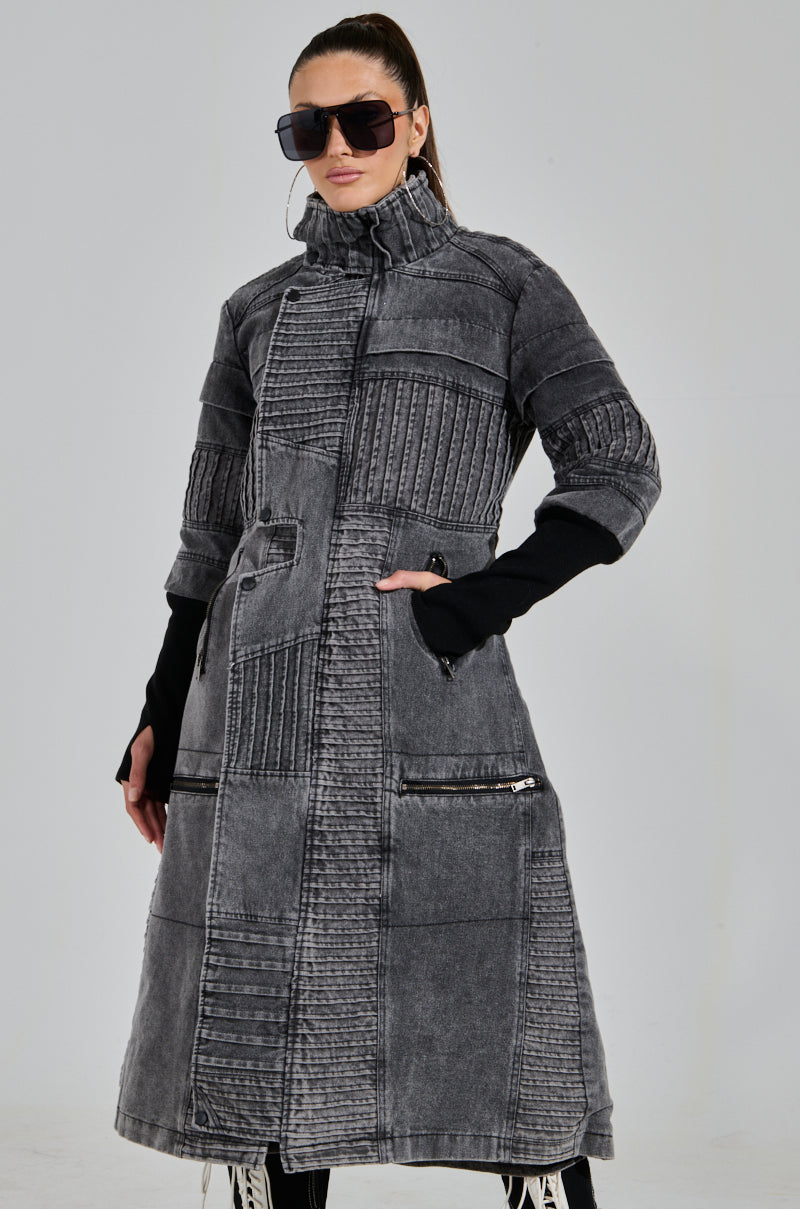 A person confidently wears the VORTEX DENIM TRENCH COAT with textured gray fabric, ribbed patchwork, zippered pockets, a high collar, and quilted accents. Sunglasses adorn their face as black sleeves peek out beneath the coat against a plain backdrop.