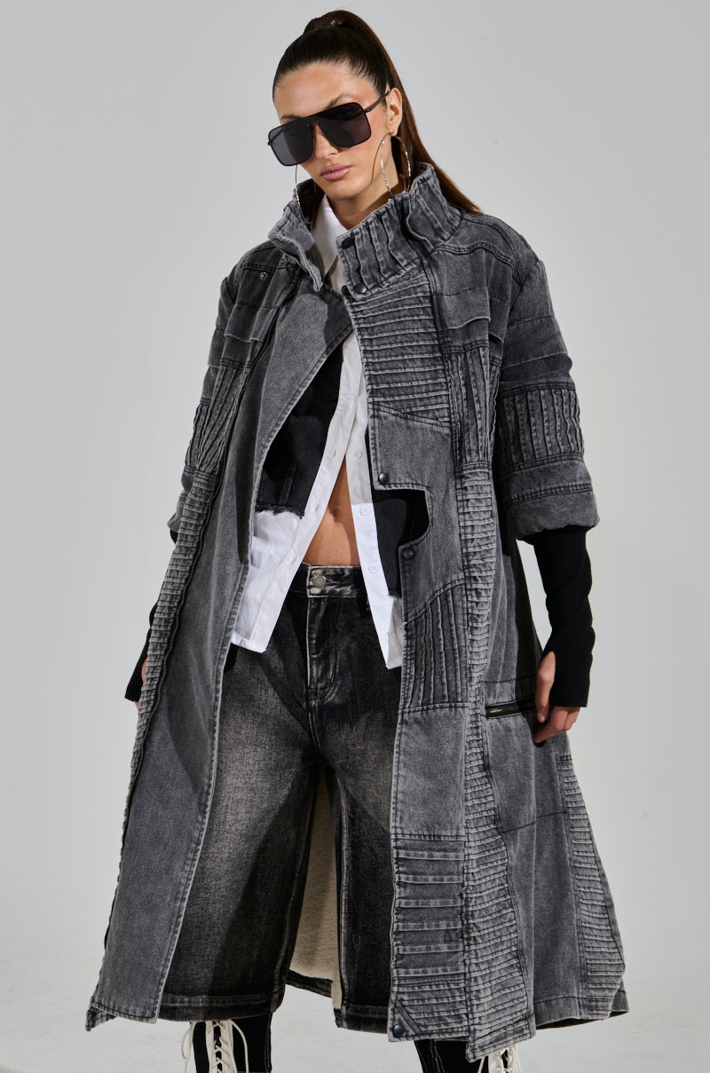 Wearing a VORTEX DENIM TRENCH COAT with textured gray ribbed patchwork, layered over a white shirt and black top. Their long hair is pulled back, complemented by large sunglasses, wide-leg dark jeans, and boots.