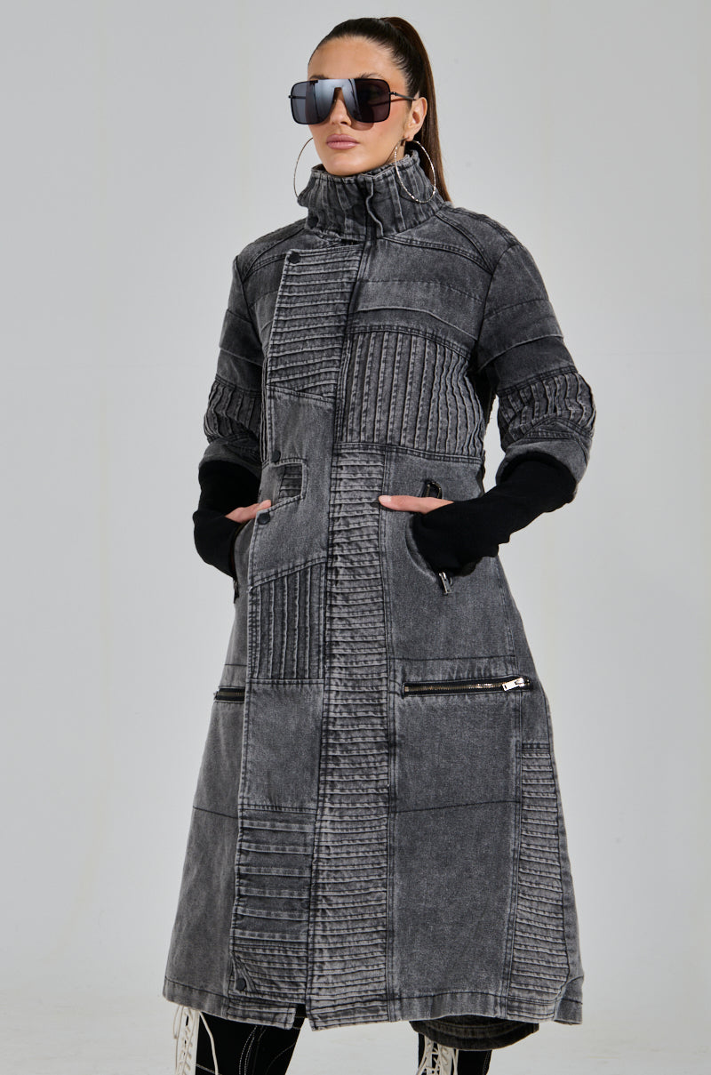 A person confidently wears the VORTEX DENIM TRENCH COAT, showcasing its ribbed patchwork, high collar, and visible zippers. They accessorize with large sunglasses and black sleeves against a light gray background.