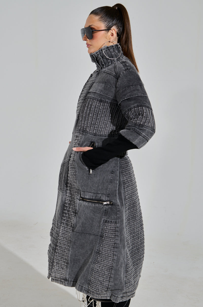 A person in a stylish VORTEX Denim Trench Coat, featuring subtle ribbed patchwork, a high collar, and pockets. Their long hair is tied back and they're wearing large sunglasses against a light-colored background.