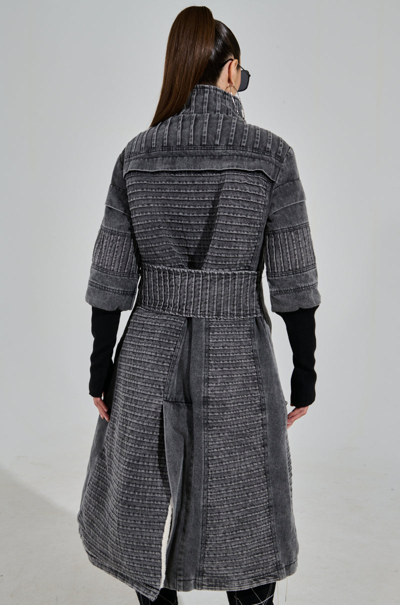 A long-haired person, wearing dark sunglasses and the VORTEX DENIM TRENCH COAT with ribbed patchwork detailing, faces away from the camera. This gray coat features a textured pattern and high collar, paired with black sleeves and leggings.