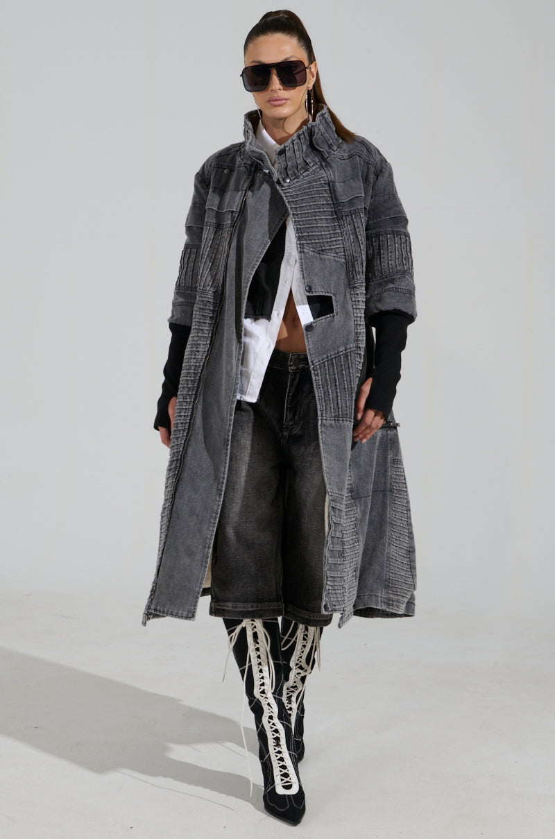 A person confidently walks in VORTEX DENIM TRENCH COAT with sherpa inner lining, sunglasses, white shirt, and black gloves. Their long boots have intricate lace details against a simple light gray background.