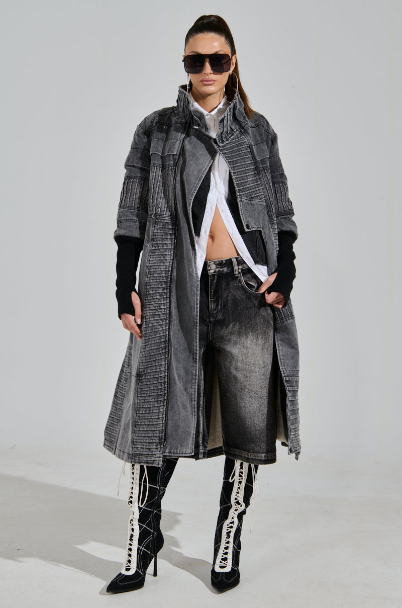 A person wearing dark oversized sunglasses and a stylish VORTEX DENIM TRENCH COAT with ribbed patchwork, a white shirt, jean shorts, long black gloves, and black knee-high boots with white laces stands against a plain background.