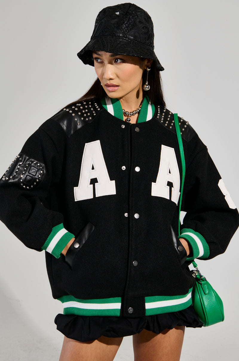 A person is wearing the ACE OF SPADES STUDDED BOMBER, a black varsity jacket with an oversized relaxed fit, featuring large white "A" letters, faux leather patches, and green accents. They have paired it with a black lace bucket hat and a green handbag. With long hair cascading down their shoulders, they are glancing to the side against a plain background.