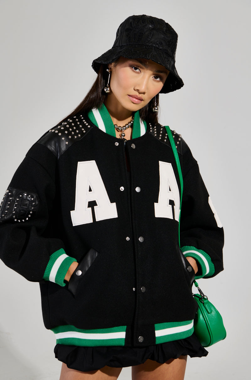 A person is wearing a black bucket hat along with the ACE OF SPADES STUDDED BOMBER jacket, which features white "A" letters and an oversized relaxed fit. The jacket is highlighted by green and white striped cuffs and hem. They are also carrying a chic green handbag.