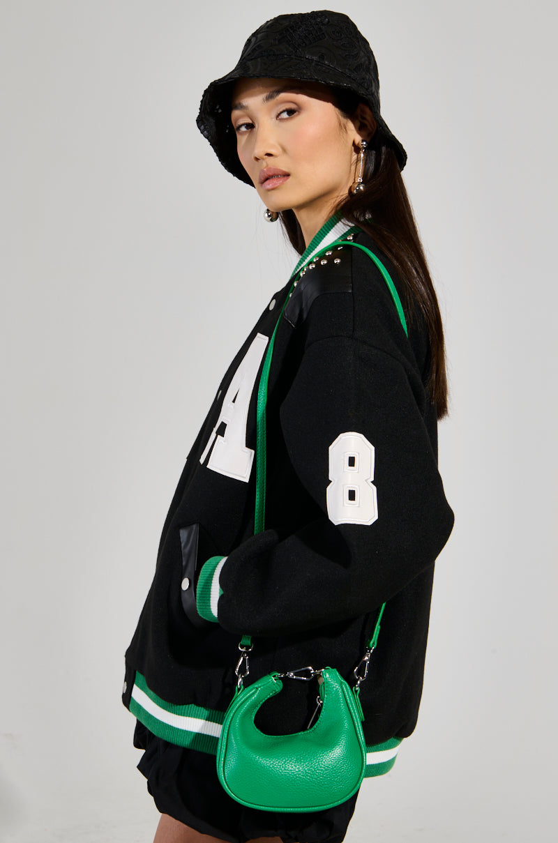 A person wearing a black bucket hat and an oversized ACE OF SPADES STUDDED BOMBER jacket with the number 8 looks to the side. The jacket, embellished with faux leather patches, showcases white lettering and green accents. They hold a green handbag against a plain backdrop.