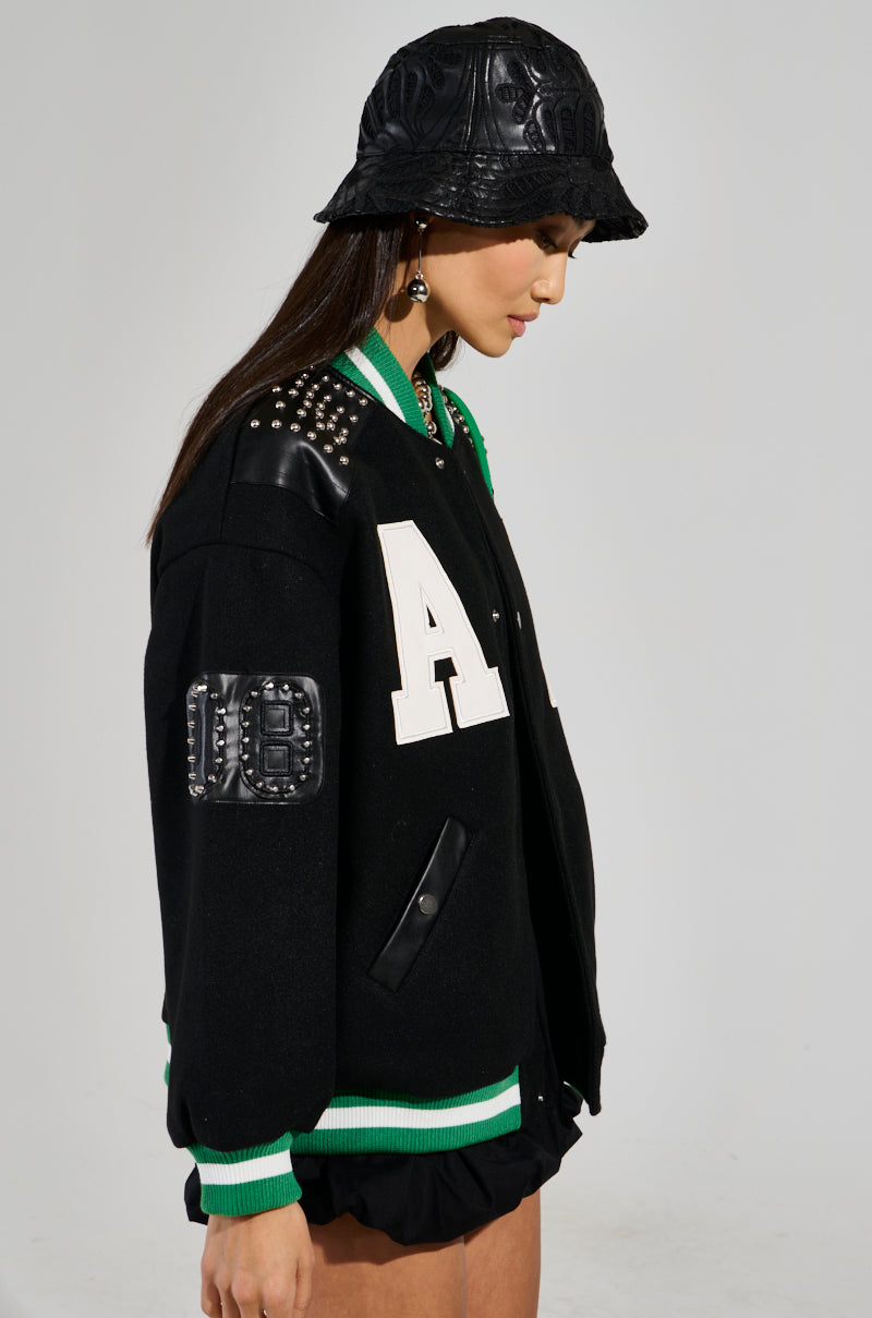 A woman wears a black bucket hat and the ACE OF SPADES STUDDED BOMBER, which showcases an oversized relaxed fit accented with faux leather patches and a striking "A" on the chest. The jacket's green and white trim pairs seamlessly with her black skirt as she gazes downward thoughtfully.