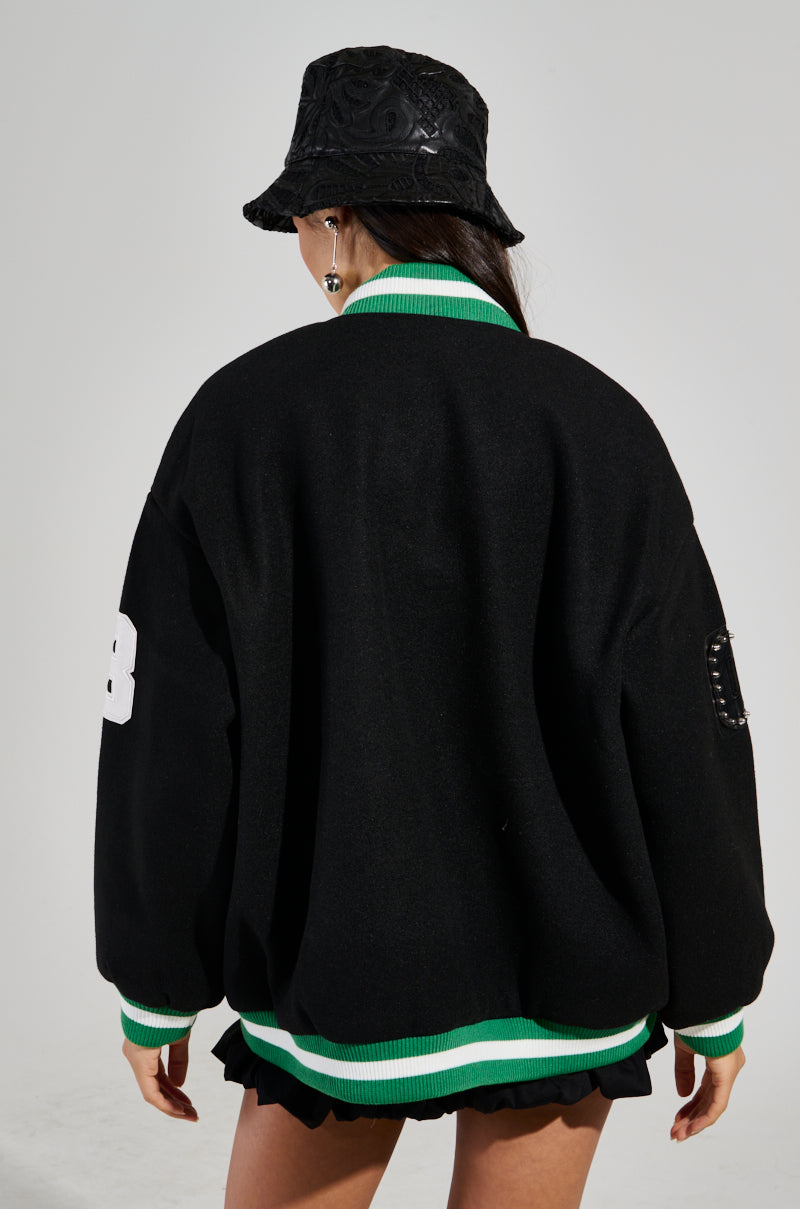 A person wearing the ACE OF SPADES STUDDED BOMBER in an oversized relaxed fit, complete with green and white trim and accented by faux leather patches, stands turned away from the camera. They pair it with a black bucket hat and black shorts featuring ruffled edges, all set against a plain background.