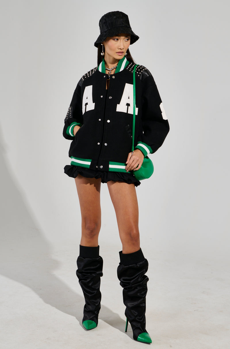A person showcases the ACE OF SPADES STUDDED BOMBER, a black varsity jacket with faux leather patches and white lettering, styled with an oversized relaxed fit. The look is completed with a black hat, a green purse, knee-high boots with green toes, a black skirt, and leg warmers against a plain white backdrop.