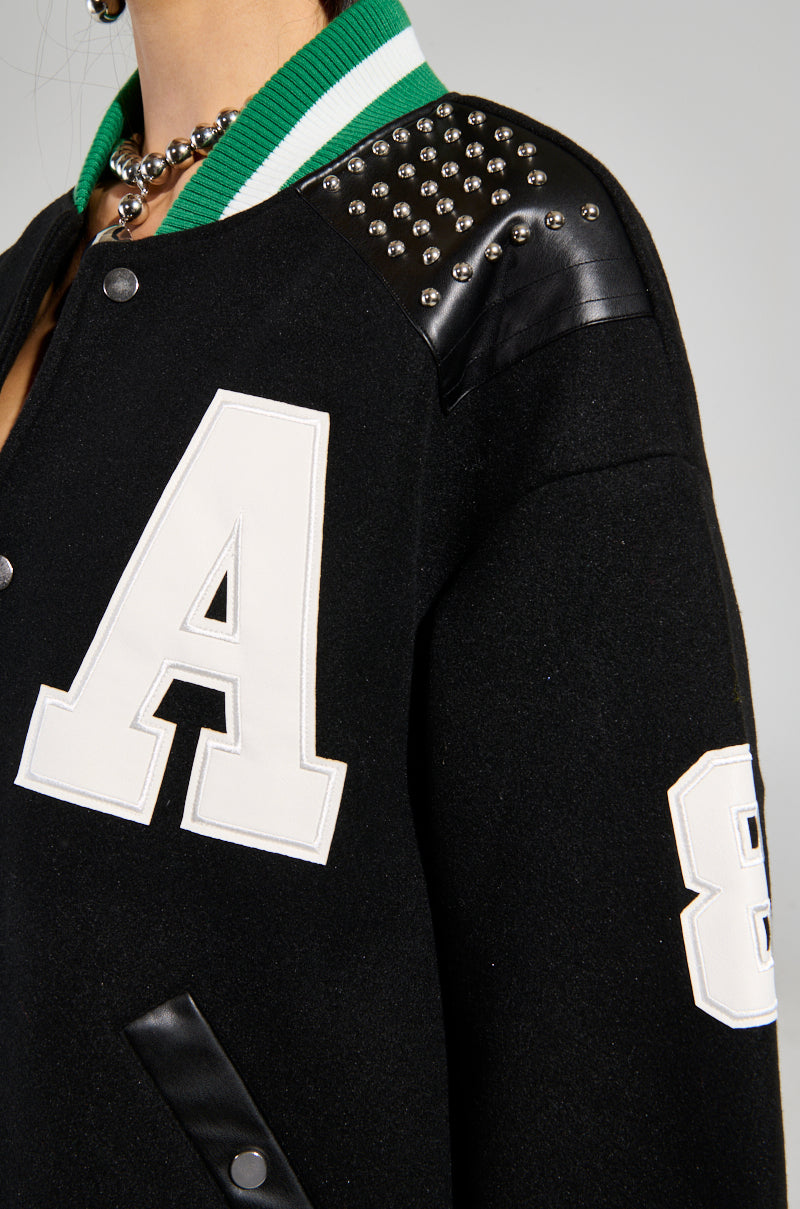 The individual is sporting the Ace of Spades Studded Bomber, an oversized black jacket adorned with a prominent white letter "A" and the number "86" on the sleeve. Its shoulders boast eye-catching faux leather patches with metal stud details, complemented by a green and white striped collar for a relaxed fit.