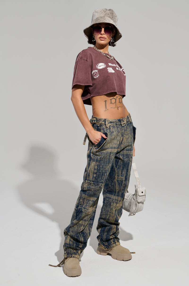 A person stands confidently wearing the NEVER LOSE ME DENIM PANT, a cropped graphic tee, and beige boots. Accessories include a bucket hat, sunglasses, and a small bag. The outfit features a mix of textures and patterns with paisley print accents that convey a trendy streetwear style.