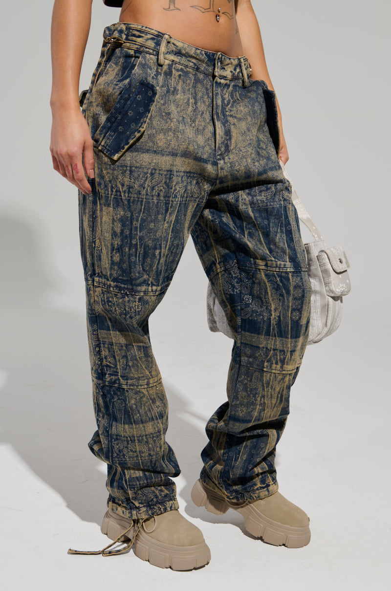 A person is wearing the NEVER LOSE ME DENIM PANT, featuring a distressed, oversized blue jeans design with a unique paisley print and two large front pockets, embodying streetwear denim style. They are carrying a light-colored bag and wearing tan chunky shoes. The image highlights the lower half of their body.
