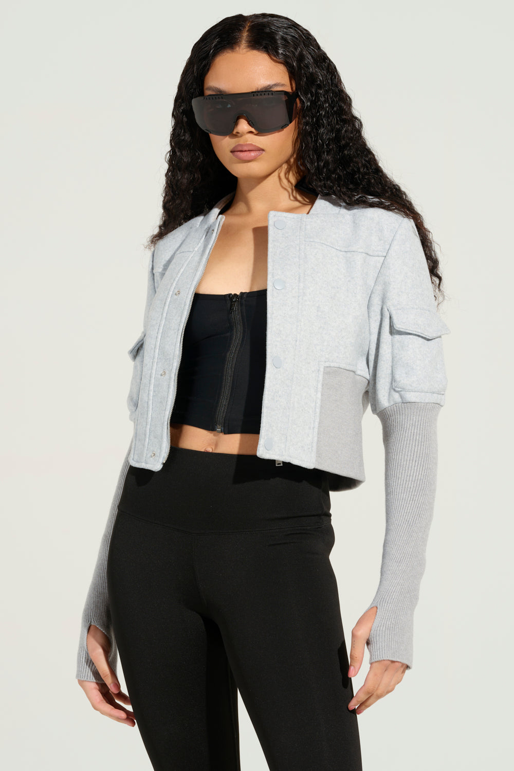 A person with long, curly hair is wearing oversized sunglasses along with the JOSIE FITTED BOMBER, which stands out due to its distinctive long sleeves. This light gray cropped jacket perfectly complements a black zip-up crop top and black leggings, all set against a simple background.