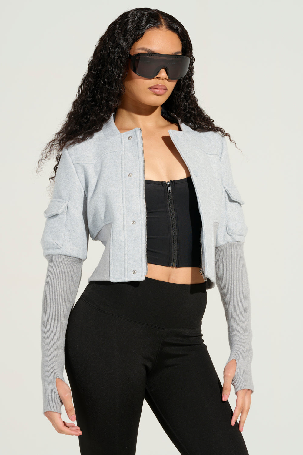 A person with long, curly hair is wearing oversized sunglasses and the JOSIE FITTED BOMBER in a light gray, cropped silhouette featuring distinctive long sleeves, paired with a black zip-up top and black pants. They are standing against a plain, light-colored background.