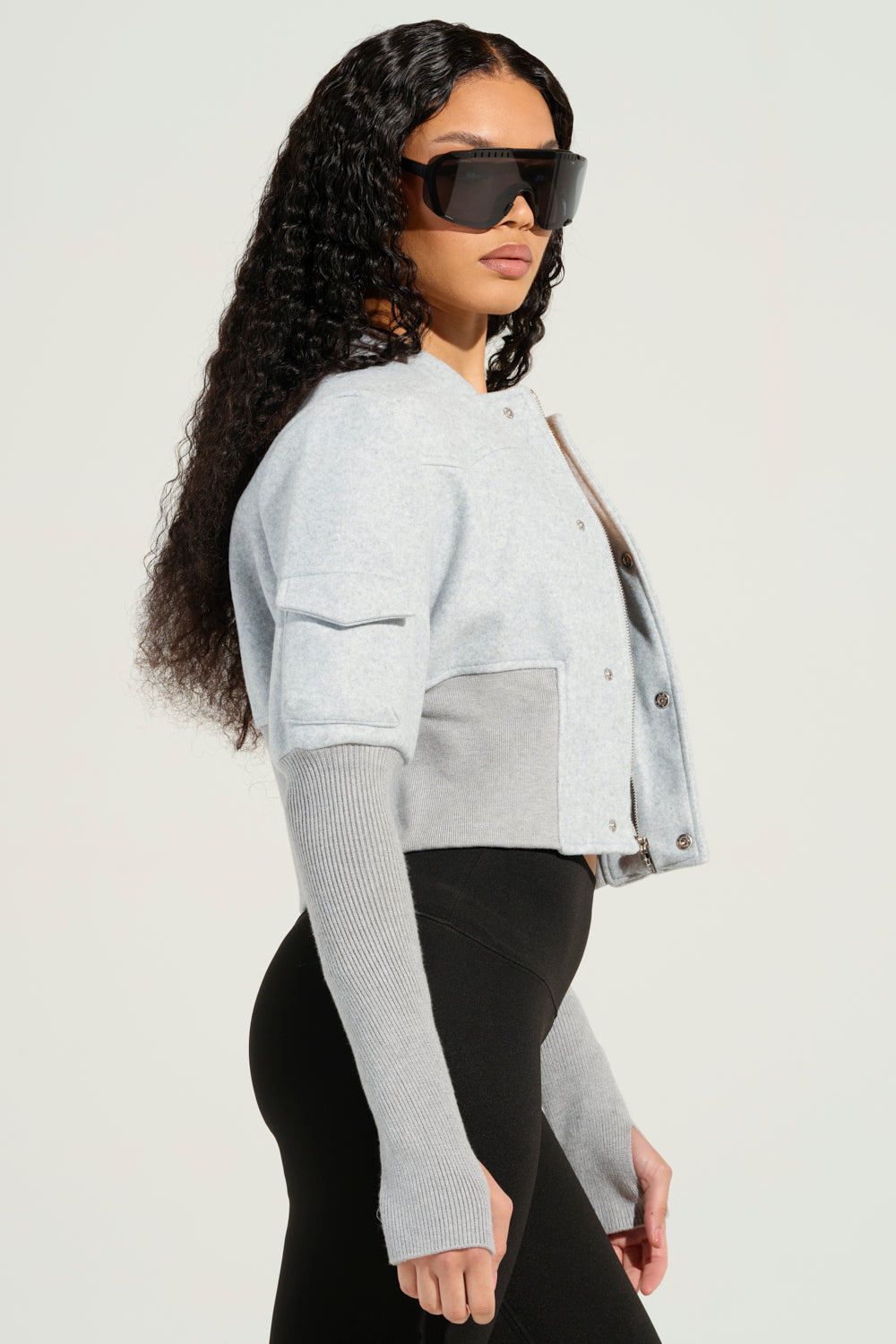 A person with long, curly hair wearing oversized sunglasses and a Josie Fitted Bomber stands sideways against a plain background. The jacket's light gray cropped silhouette pairs perfectly with black leggings, and the individual has their hands tucked into its distinctive long sleeves.