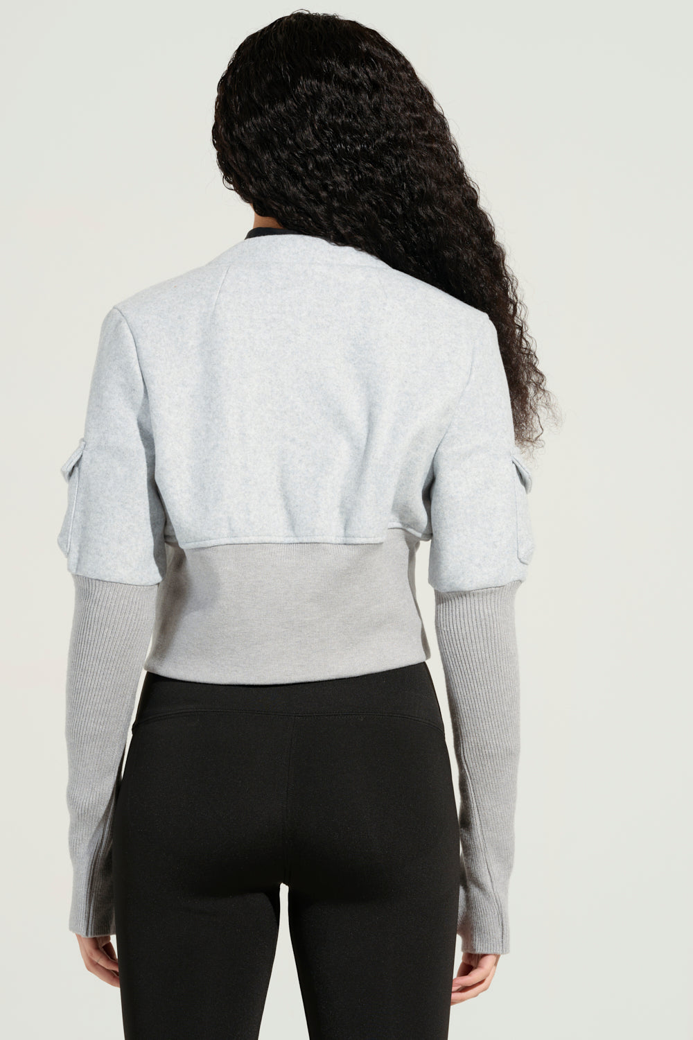A person with long curly hair is seen from behind, dressed in the JOSIE FITTED BOMBER featuring a light gray cropped silhouette, paired with black leggings. The bomber's distinct long sleeves are ribbed, all set against a plain light background.