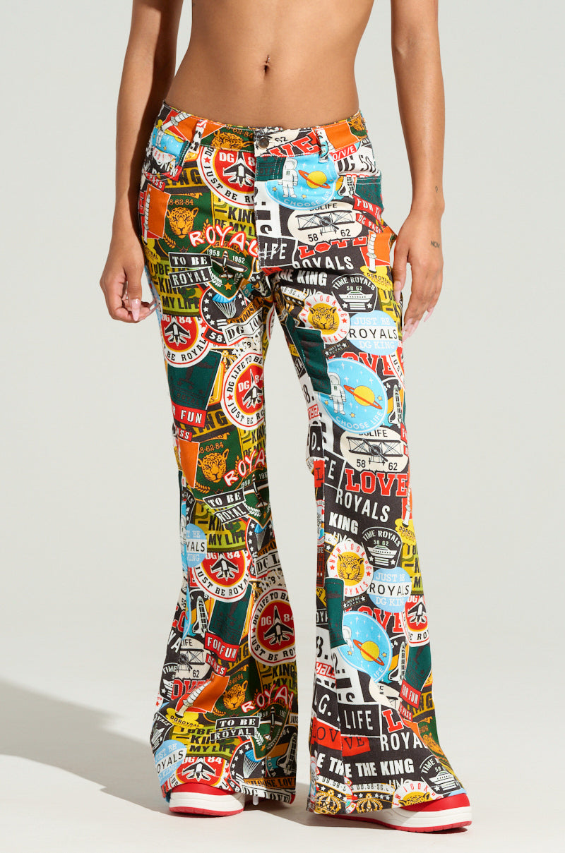 A person models the RED LIGHT LOW RISE FLARE DENIM featuring vibrant, aviation-inspired graphic prints with various logos, symbols, and text. These colorful pants showcase a retro and playful design. The model's midriff is visible as they pair the look with red and white shoes.