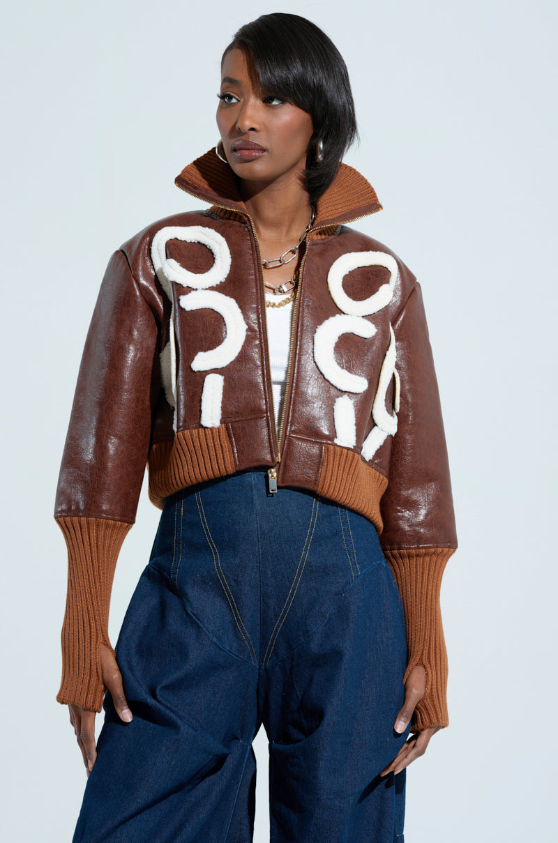 A person poses confidently wearing the GIULIA SHERPA TRIM BOMBER, a brown faux leather jacket featuring bold white circular patterns and sherpa trim over a knit collar and cuffs. They style it with trendy high-waisted blue pants and layered necklaces against a plain background.