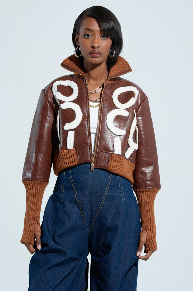 A person dressed in the GIULIA SHERPA TRIM BOMBER, featuring a brown faux leather design with white circular patterns and sherpa detailing, paired with high-waisted blue jeans. They have short, styled hair and are accessorized with hoop earrings and a gold chain necklace.