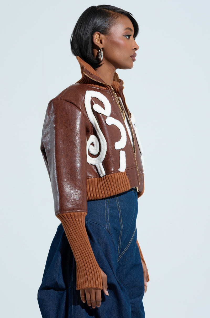 A woman in the GIULIA SHERPA TRIM BOMBER jacket, featuring a brown exterior with white sherpa trim and long ribbed sleeves, paired with high-waisted blue jeans. She stands sideways, looking forward, with short black hair and hoop earrings.