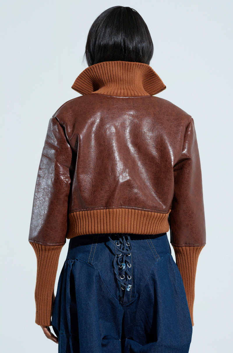 A person with dark hair is shown from behind, wearing the GIULIA SHERPA TRIM BOMBER, featuring a high ribbed collar and long ribbed cuffs that complement its brown faux leather finish. They are paired with blue pants featuring laces on the back. The plain light gray background enhances the stylish look of the outfit.