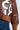 Close-up of a person wearing the GIULIA SHERPA TRIM BOMBER, a brown faux leather jacket adorned with white textured letters. This stylish piece features sherpa trim for added warmth and includes ribbed cuffs and waistband. The look is complemented by blue denim pants with visible yellow stitching.